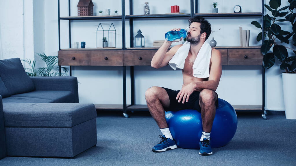κουρασμένος αθλητής με πετσέτα κάθεται σε μπάλα γυμναστικής και πόσιμο νερό από αθλητικό μπουκάλι - Φωτογραφία, εικόνα