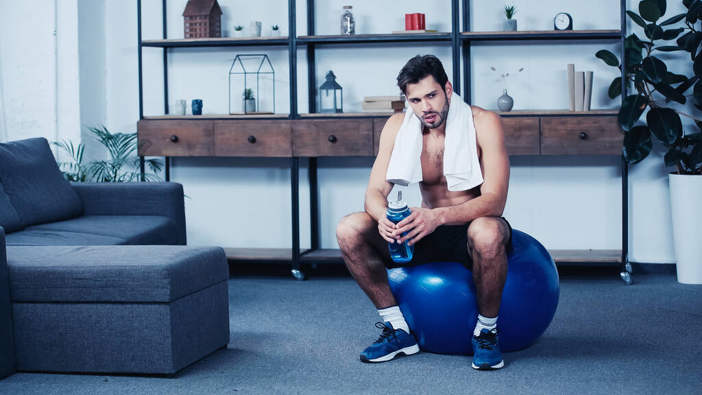 Erschöpfter Sportler mit Handtuch sitzt auf Fitnessball mit Sportflasche  - Foto, Bild