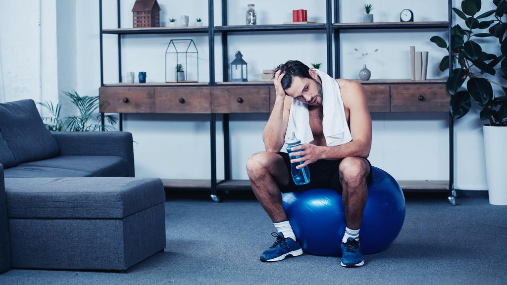 Müder Sportler mit Handtuch sitzt auf Fitnessball mit Sportflasche - Foto, Bild