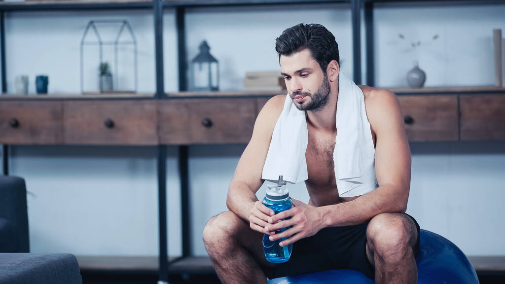 sportif épuisé avec une serviette assise sur une balle de fitness et tenant une bouteille de sport  - Photo, image