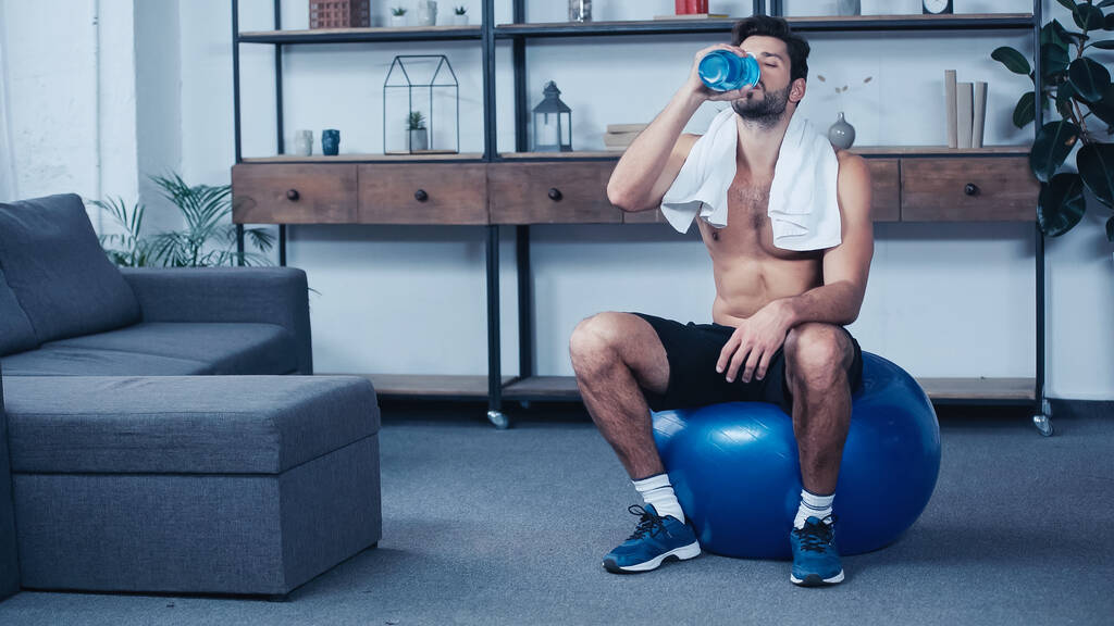 sportif fatigué de toute la longueur avec une serviette assise sur une balle de fitness et tenant une bouteille de sport tout en buvant de l'eau - Photo, image