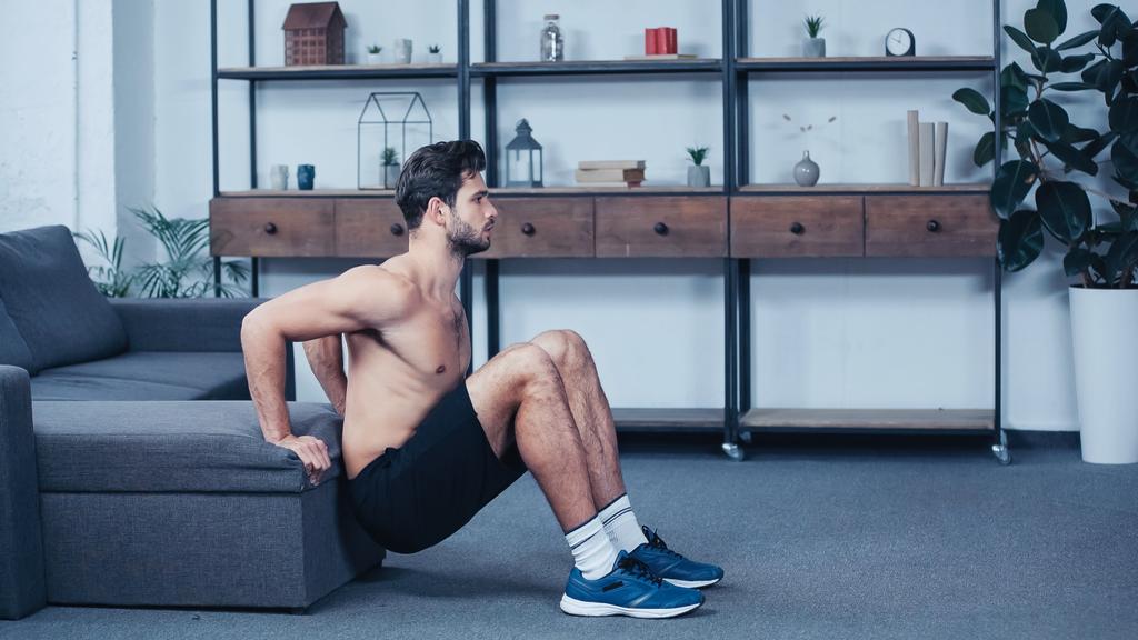 side view of shirtless sportsman in shorts squatting near sofa at home - Foto, Bild