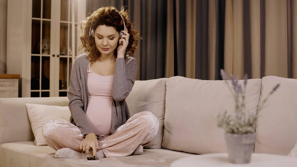 Беременная женщина в наушниках с помощью смартфона на диване дома  - Фото, изображение