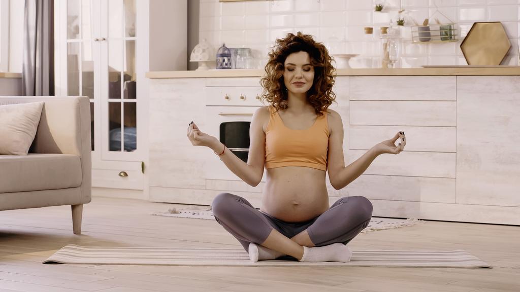 Mujer embarazada sonriente con los ojos cerrados sentada en postura de yoga en casa  - Foto, imagen