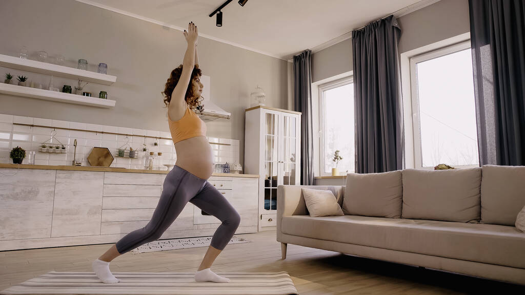 Вид сбоку на тренировку беременной женщины на фитнес-коврике возле дивана дома  - Фото, изображение