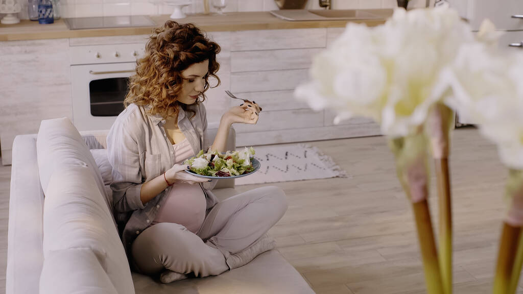 Göndör terhes nő kezében saláta és villa a kanapén otthon  - Fotó, kép