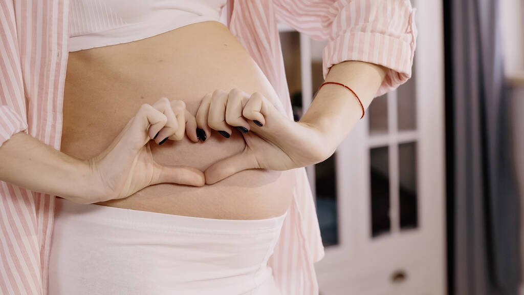 Oříznutý pohled na těhotné ženy ukazující srdeční znamení v blízkosti břicha doma  - Fotografie, Obrázek