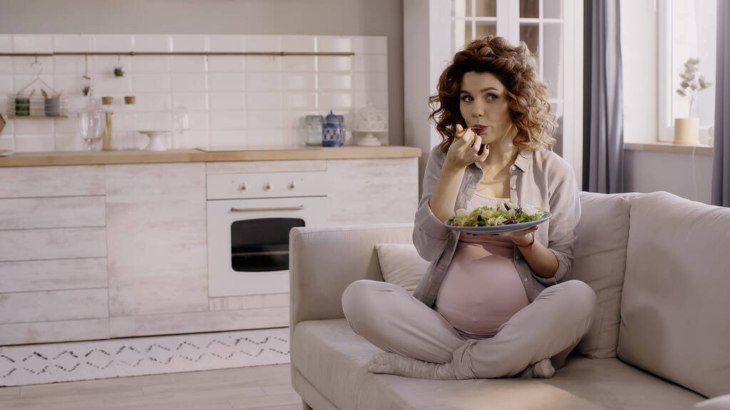 Kıvırcık hamile kadın evdeki koltukta taze salata yiyor.  - Fotoğraf, Görsel