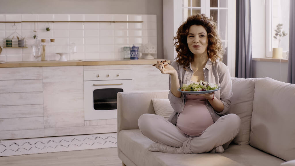 Boldog terhes nő kezében tányér friss saláta és villa a kanapén  - Fotó, kép