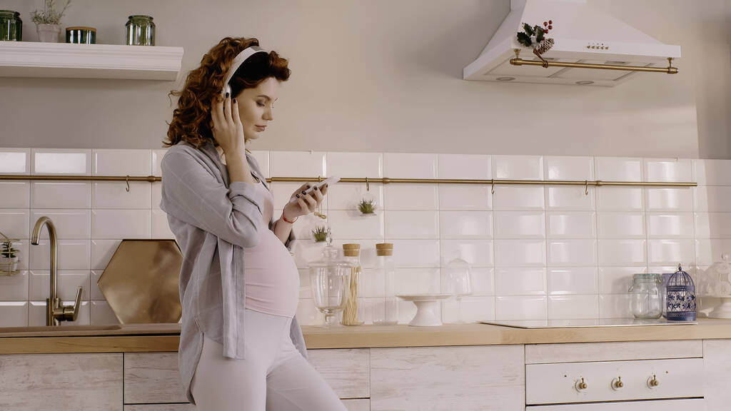 Νεαρή έγκυος γυναίκα ακούει μουσική στα ακουστικά και κρατώντας smartphone στην κουζίνα  - Φωτογραφία, εικόνα