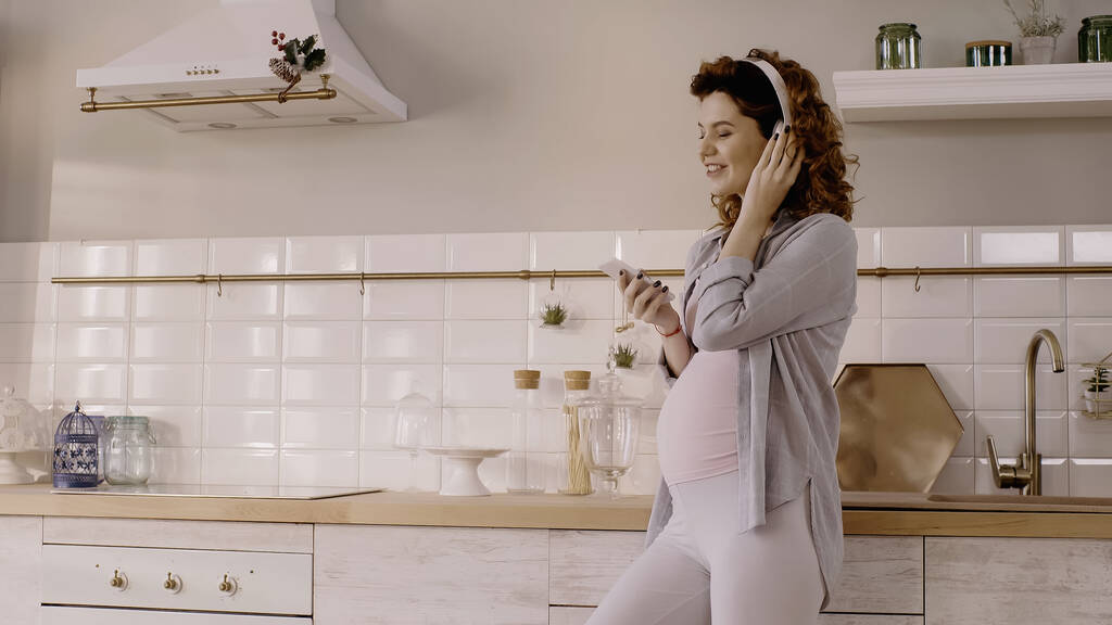 Χαρούμενη έγκυος γυναίκα που χρησιμοποιεί ακουστικά και κινητό τηλέφωνο στην κουζίνα  - Φωτογραφία, εικόνα