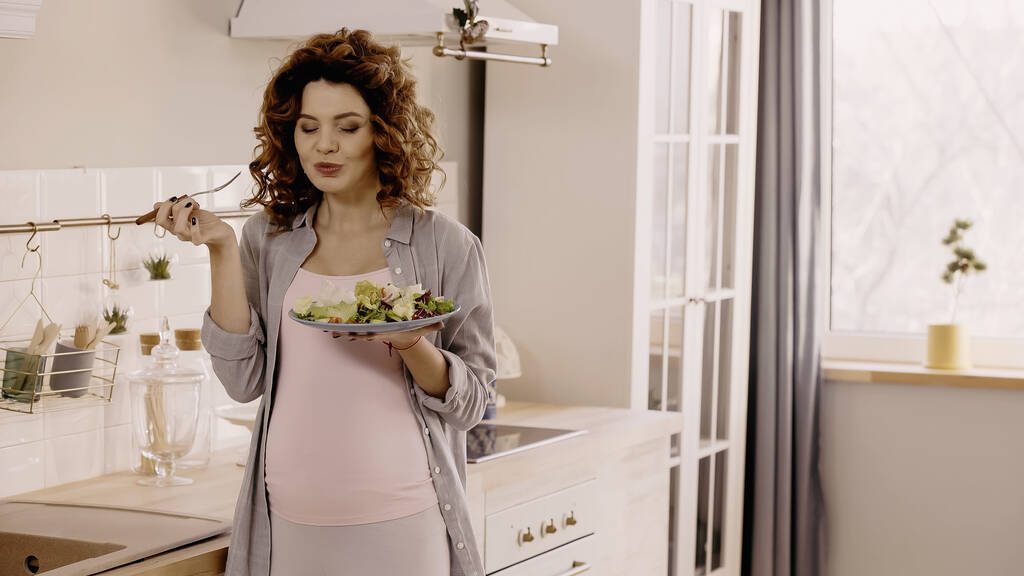 Fröhliche Schwangere hält frischen Salat und Gabel in der Küche  - Foto, Bild