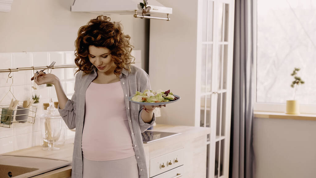 Σγουρή έγκυος γυναίκα κρατώντας πιρούνι και σαλάτα, ενώ κοιτάζοντας την κοιλιά στην κουζίνα  - Φωτογραφία, εικόνα