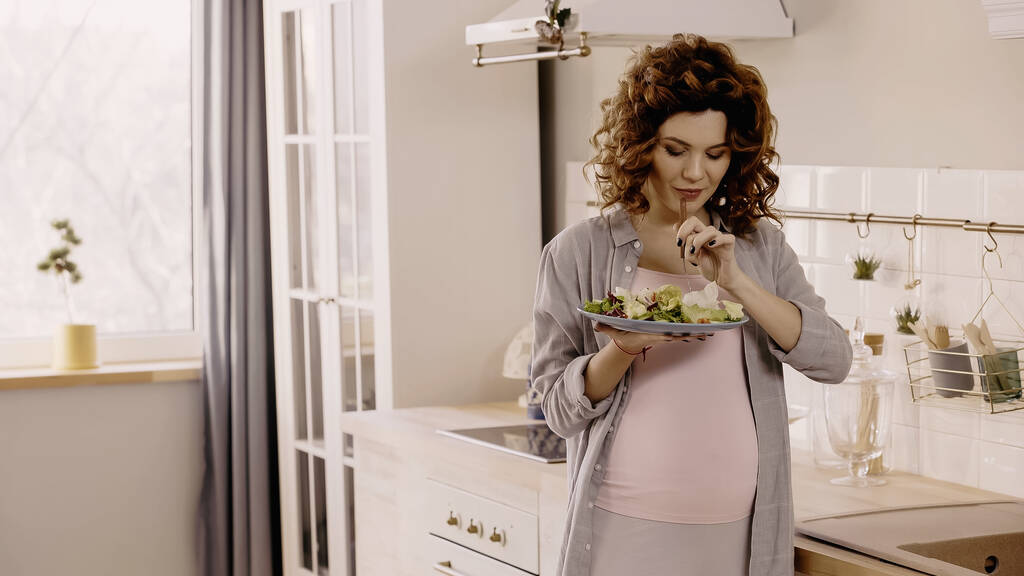 Young pregnant woman holding fresh salad and fork in kitchen  - Foto, Bild