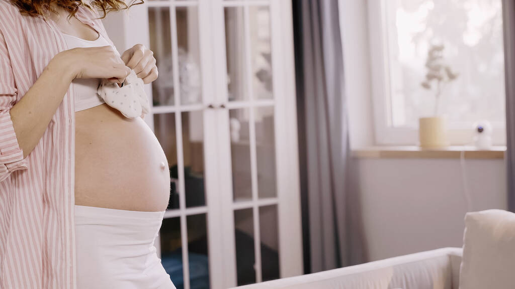 Vágott kilátás a terhes nő gazdaság baba cipő a nappaliban  - Fotó, kép