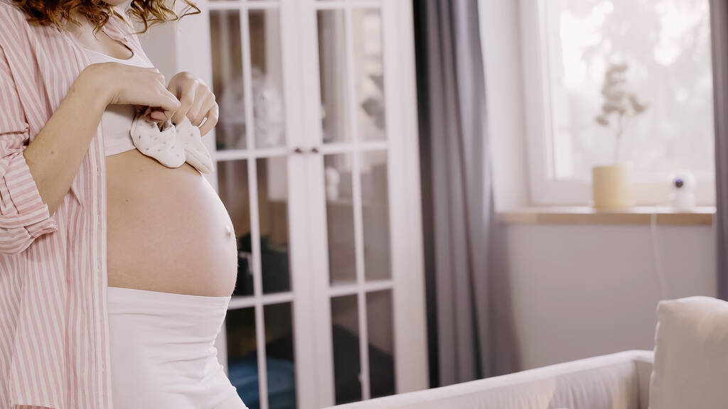 Vista cortada da mulher grávida encaracolada segurando botas de bebê perto da barriga em casa  - Foto, Imagem