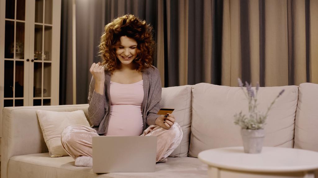Femme enceinte excitée tenant une carte de crédit et montrant un geste oui lors des achats en ligne à la maison  - Photo, image