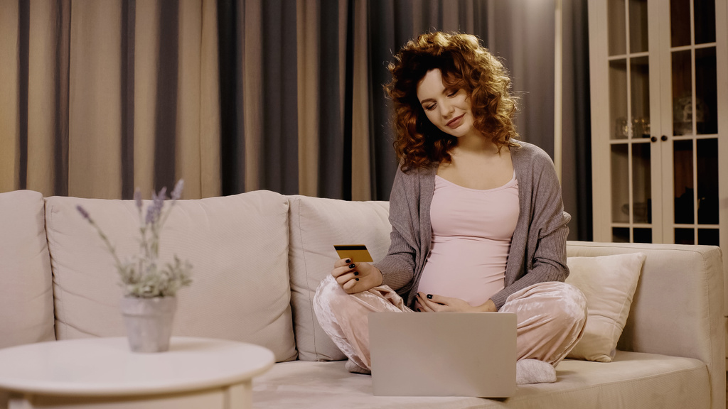 Mujer embarazada sosteniendo la tarjeta de crédito cerca del ordenador portátil y la planta borrosa en el sofá en casa  - Foto, imagen