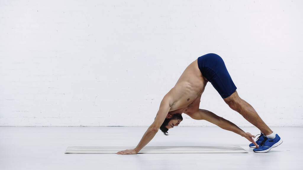 side view of shirtless sportsman stretching back on fitness mat near white brick wall - Fotó, kép