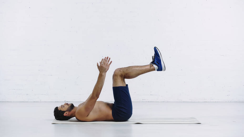 zijaanzicht van shirtloze sportman in korte broek liggend op de rug en aan het trainen op fitness mat in de buurt van witte bakstenen muur - Foto, afbeelding