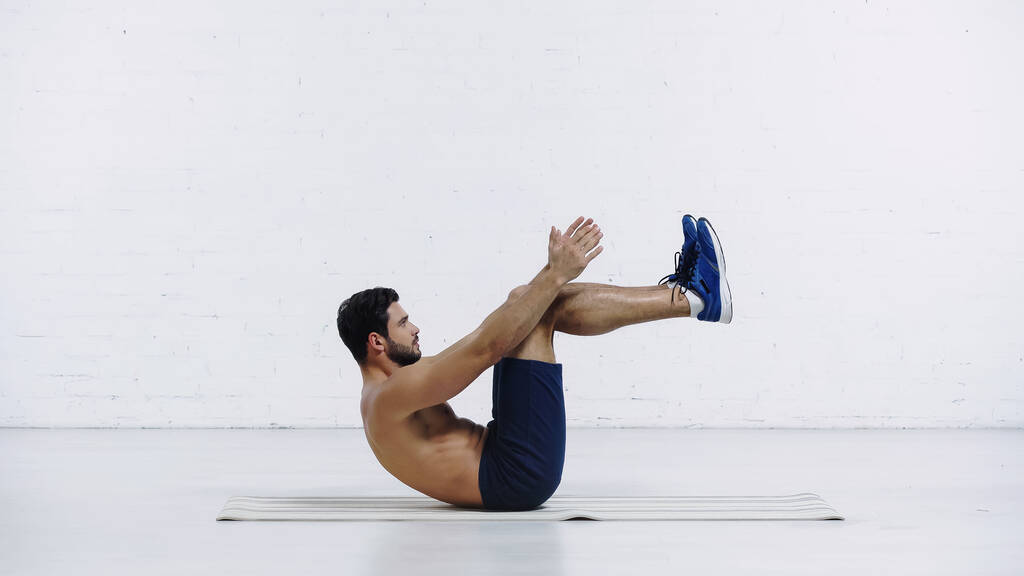 zijaanzicht van een bebaarde sportman in korte broek die aan het trainen is op een fitnessmat in de buurt van een witte bakstenen muur - Foto, afbeelding