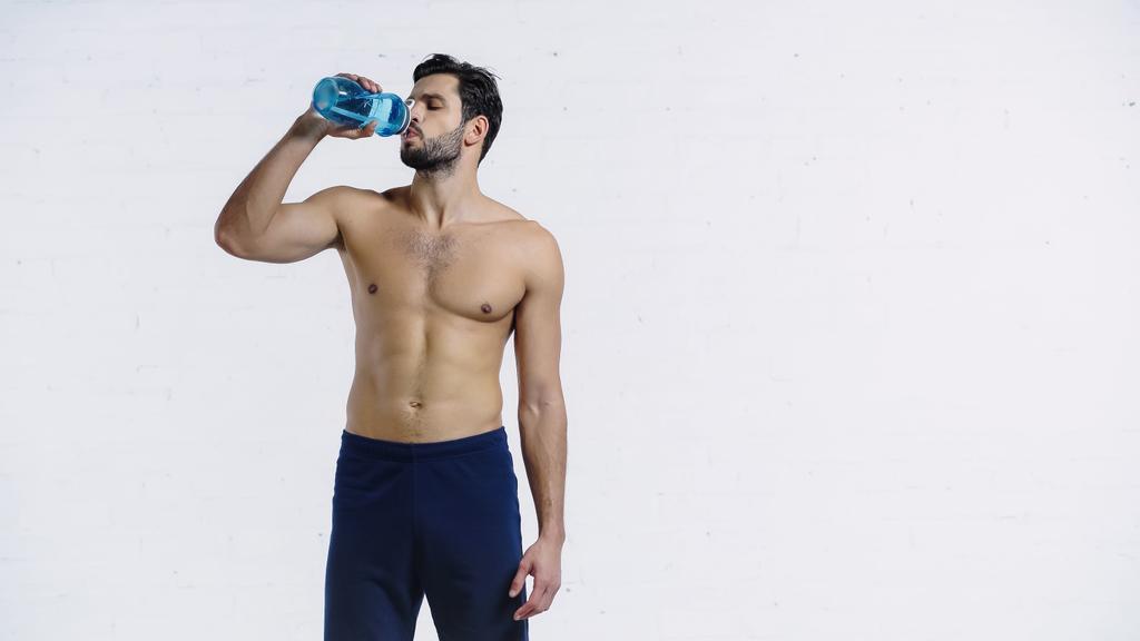 bearded sportsman in shorts holding blue sports bottle and drinking water near white brick wall  - Photo, Image