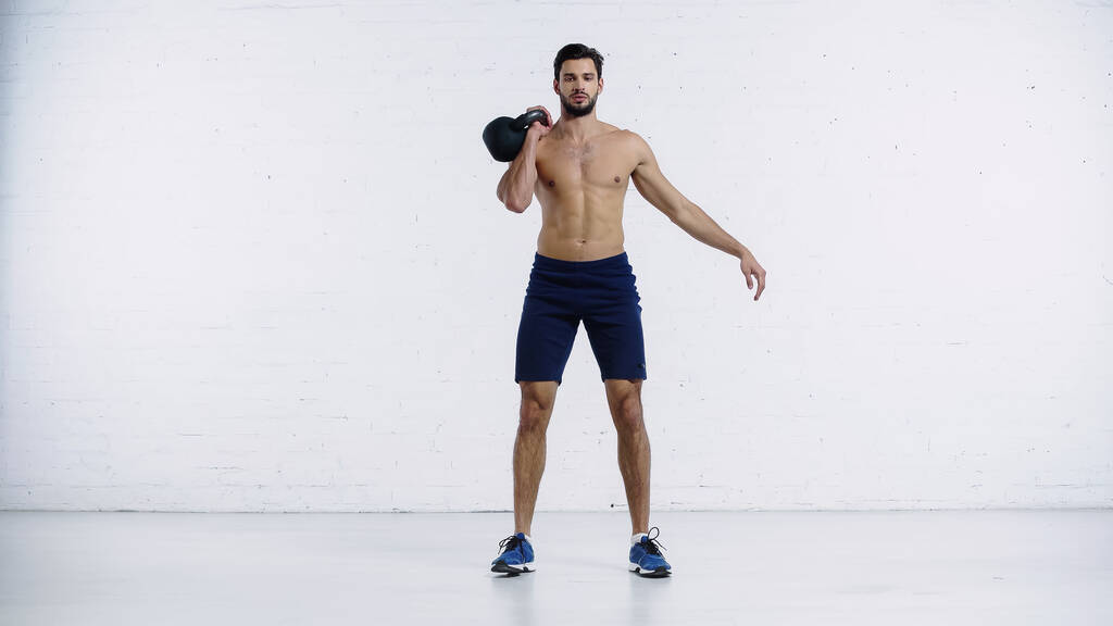 Мужчина без рубашки и спортивные упражнения с тяжелыми гирями возле стены из белого кирпича  - Фото, изображение