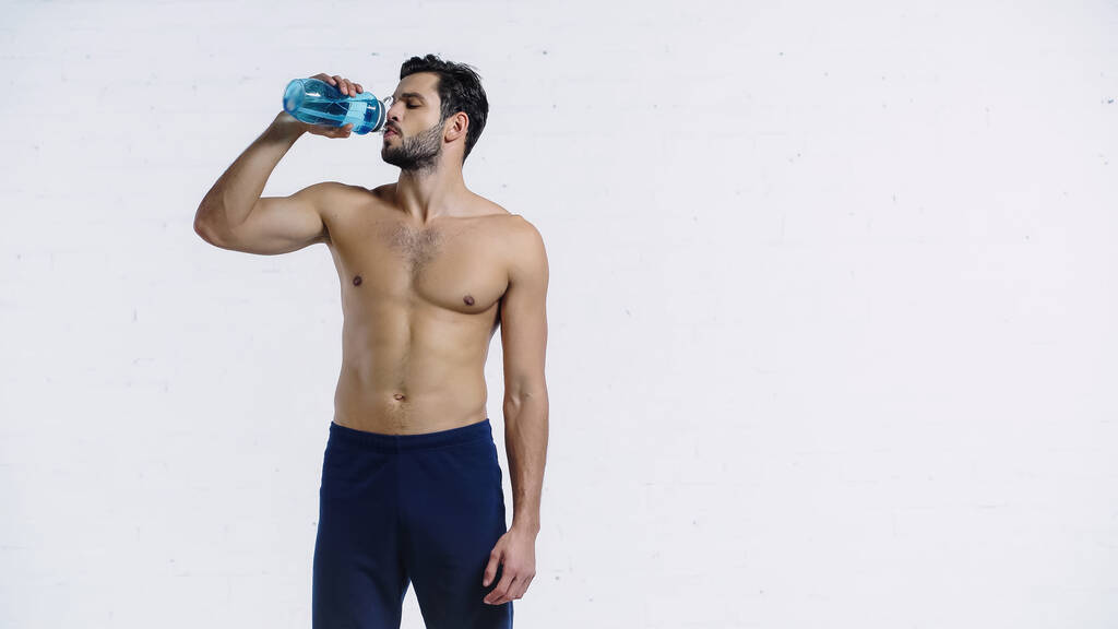 μυώδης αθλητής σε μπλε σορτς πόσιμο νερό από αθλητικό μπουκάλι κοντά σε λευκό τούβλο τοίχο  - Φωτογραφία, εικόνα