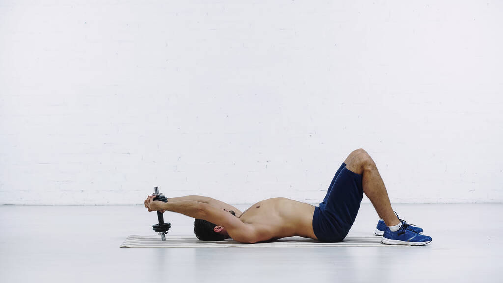 side view of shirless sportsman in shorts training with heavy dumbbell while lying on fitness mat near white brick wall - Zdjęcie, obraz
