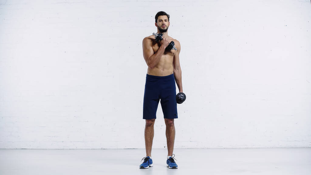 volledige lengte van sportieve man in shorts en sneakers uit te werken met halters op wit  - Foto, afbeelding