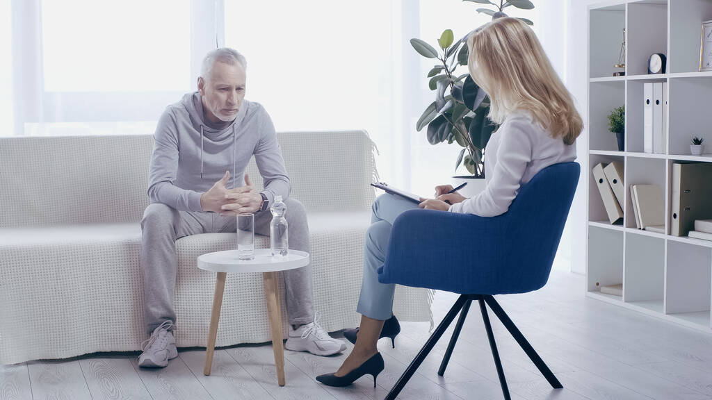 volwassen sportman en blond psycholoog met gesprek in consulting room - Foto, afbeelding