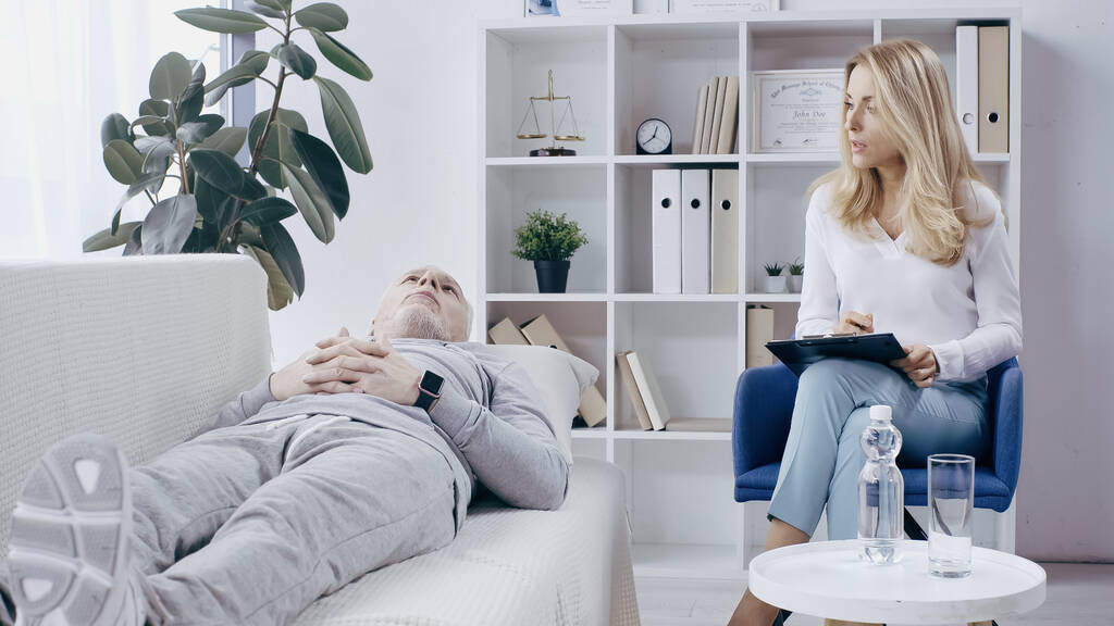psychothérapeute blonde avec presse-papiers parlant à l'homme déprimé en vêtements de sport couché sur le canapé dans la salle de consultation  - Photo, image