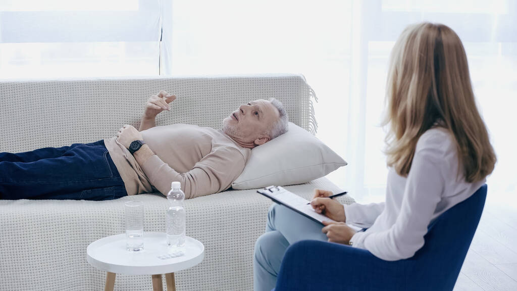 psychothérapeute blonde tenant stylo près du presse-papiers et l'écoute homme mature couché sur le canapé dans la salle de consultation - Photo, image