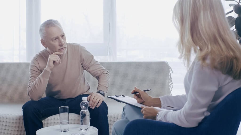 blond psycholoog houden klembord in de buurt pensive middelbare leeftijd man in consulting kamer  - Foto, afbeelding