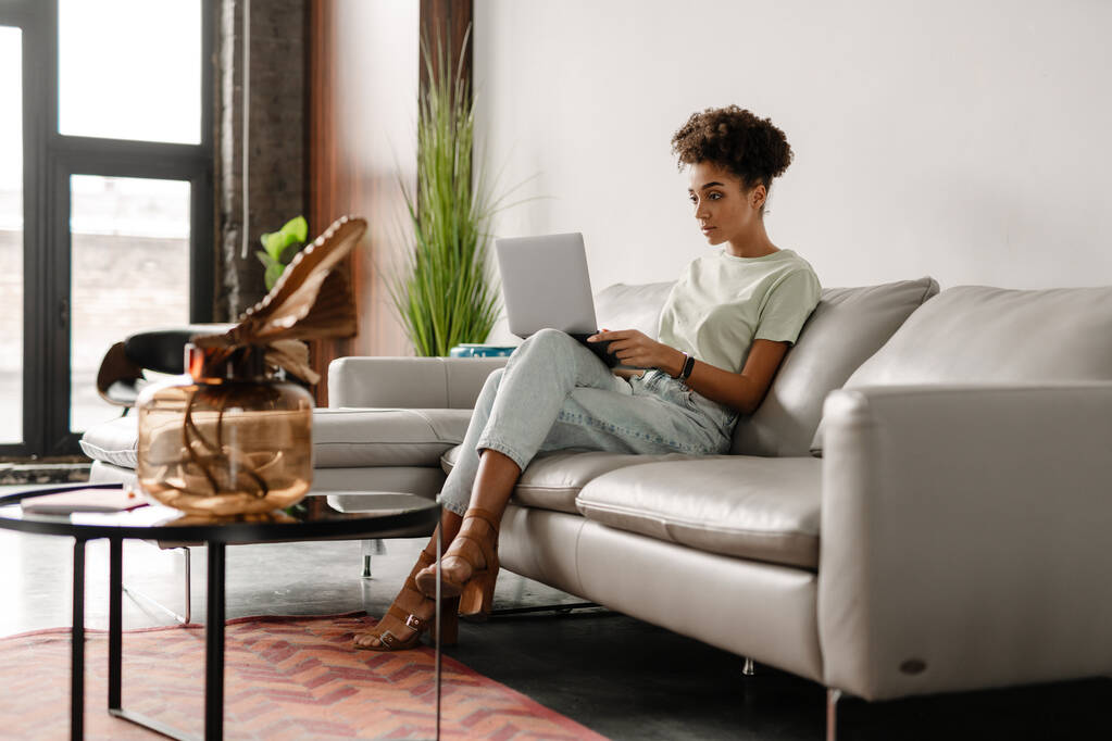 Молода чорна жінка працює з ноутбуком, сидячи на дивані вдома
 - Фото, зображення