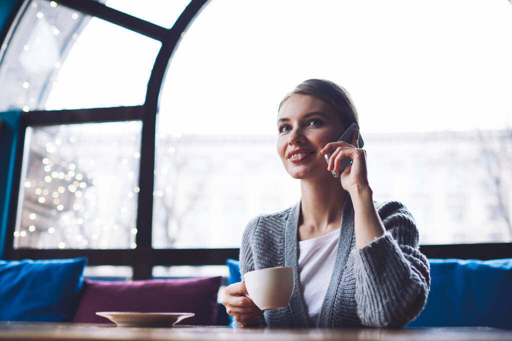 Alacsony szögben mosolygós nő alkalmi ruhát néz félre, miközben ül az asztalnál a kávézó csésze forró kávé és a beszélgetés okostelefonon - Fotó, kép