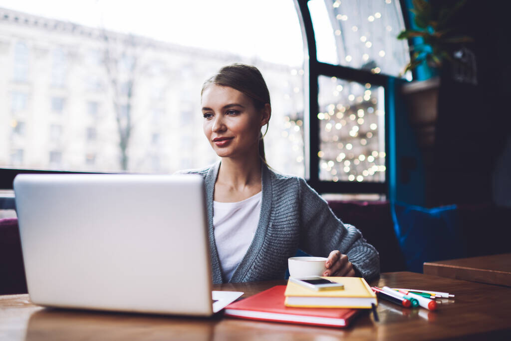 Dreamy kobieta freelancer w casual ubrania patrząc daleko siedząc przy stole z filiżanką kawy i pracy na nowoczesnym laptopie - Zdjęcie, obraz