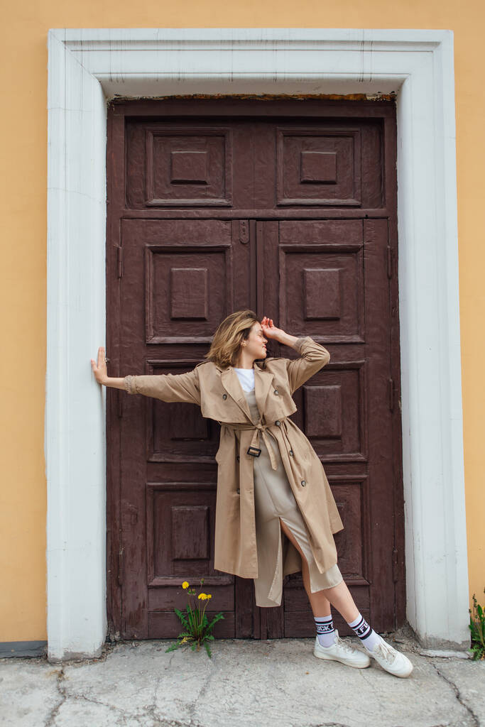 Joven mujer milenaria sonriente con pelo salvaje vestida con un abrigo de otoño posando cerca de la puerta de un viejo edificio. - Foto, Imagen