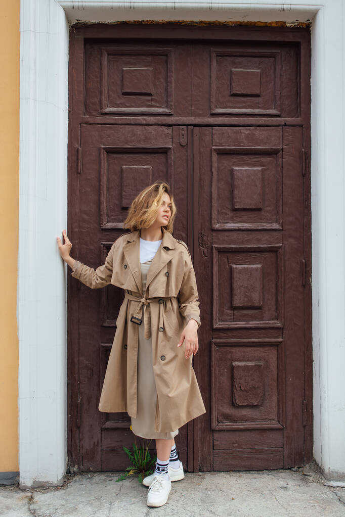 Junge lächelnde Millennial-Frau mit wilden Haaren im Herbstmantel posiert vor der Tür eines alten Gebäudes. - Foto, Bild