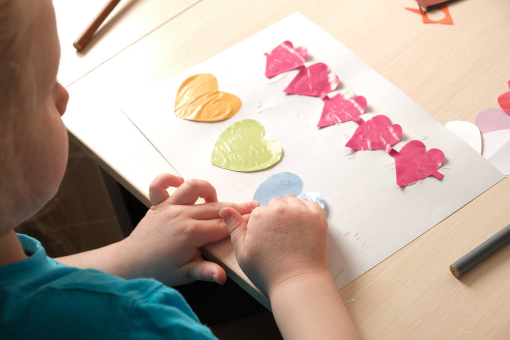 Przedszkolanka w niebieskiej koszulce sprawia, że aplikacja siedzi przy stole, serca wycięte z kolorowych bukietów na kartkę na Walentynki, kreatywność dzieci, atopowe zapalenie skóry na rękach dziecka - Zdjęcie, obraz
