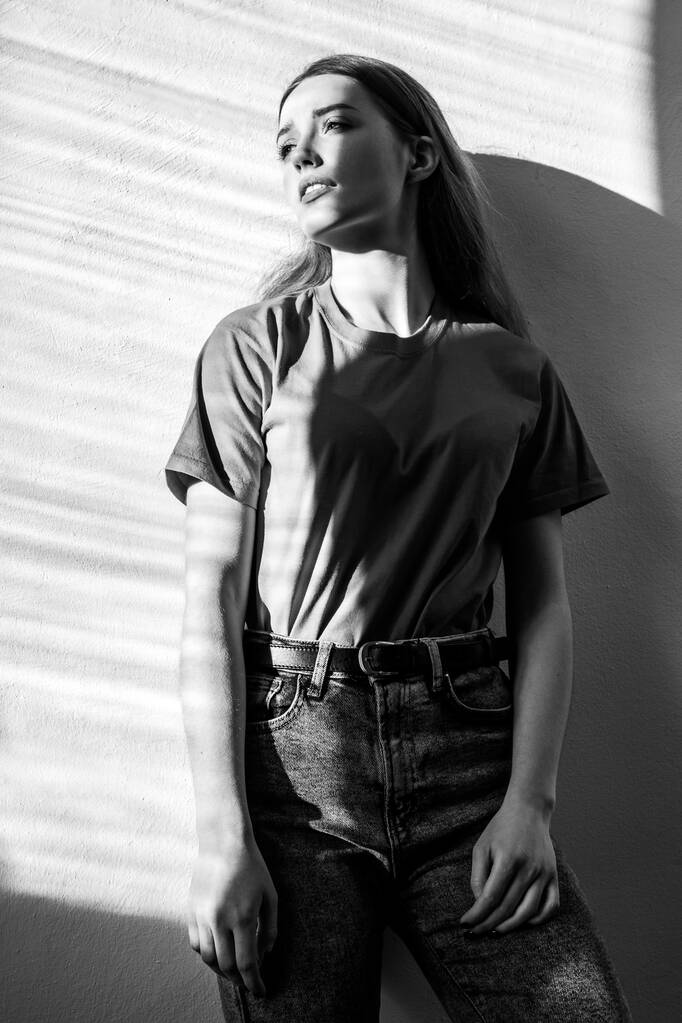 Jeune adulte belle femme portant T-shirt et jeans debout regardant loin avec expression faciale romantique coûteuse. Photographie en noir et blanc, prise de vue d'intérieur éclairée par la lumière du soleil depuis la fenêtre - Photo, image