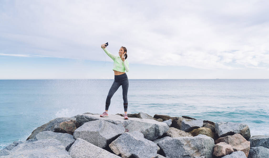 Onnellinen nuori nainen urheiluvaatteissa seisoo kivellä ja ottaa selfie rannikolla samalla heiluttaen käsi ja tervehdys ystävä matkapuhelimeen - Valokuva, kuva