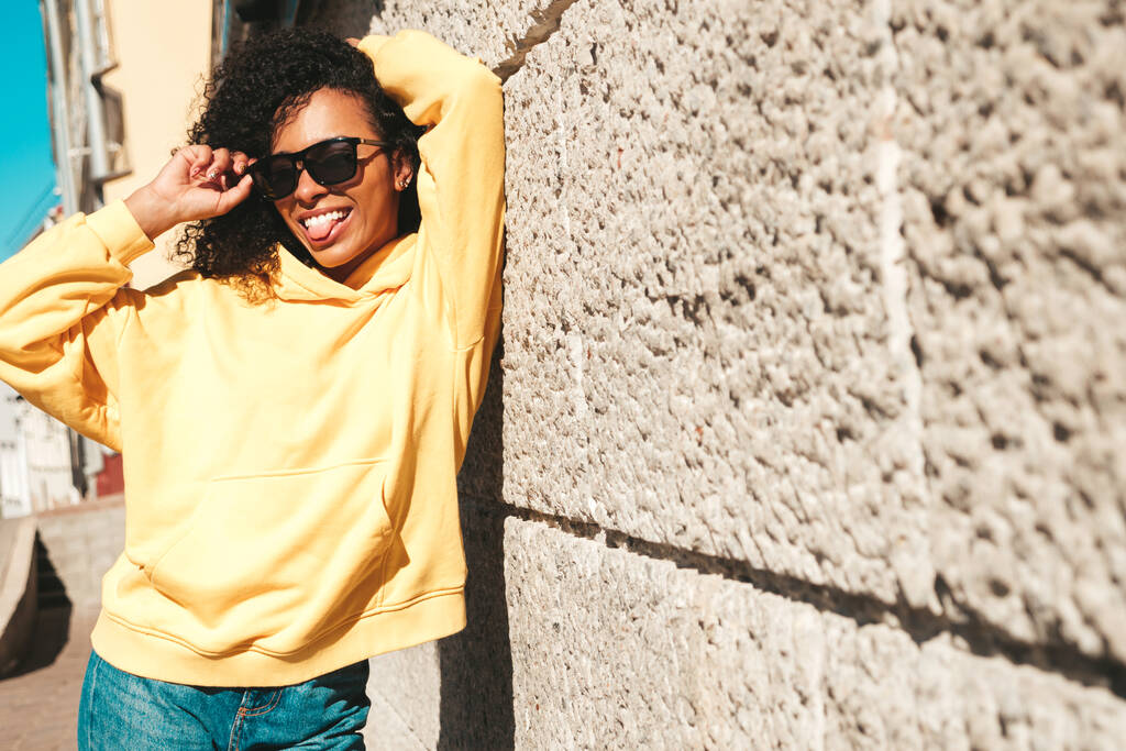 Schöne Frau mit Afro-Locken-Frisur. Lächelndes Hipster-Model in gelbem Kapuzenpulli. Sexy unbeschwerte Frau posiert mit Sonnenbrille auf der Straße. Fröhlich und glücklich - Foto, Bild