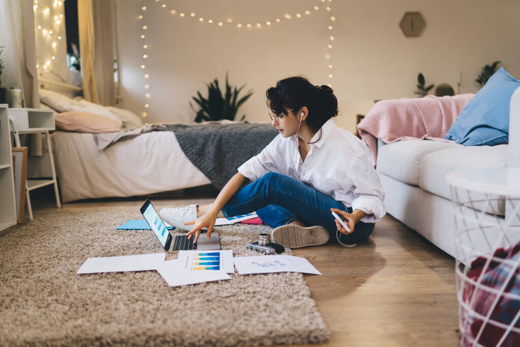 Side view koko kehon positiivinen nuori naispuolinen opiskelija tai freelancer työskentelee netbook ja kuunnella musiikkia kuulokkeiden istuessaan lattialla paperit kodikas makuuhuone - Valokuva, kuva