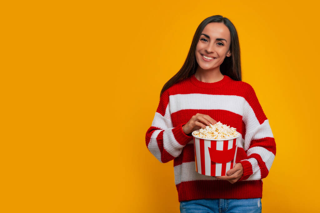 Close up portrait of young beautiful brunette woman in sweater while she holds big bucket of popcorn for watching movie in cinema and emotionally looking on camera - Photo, image