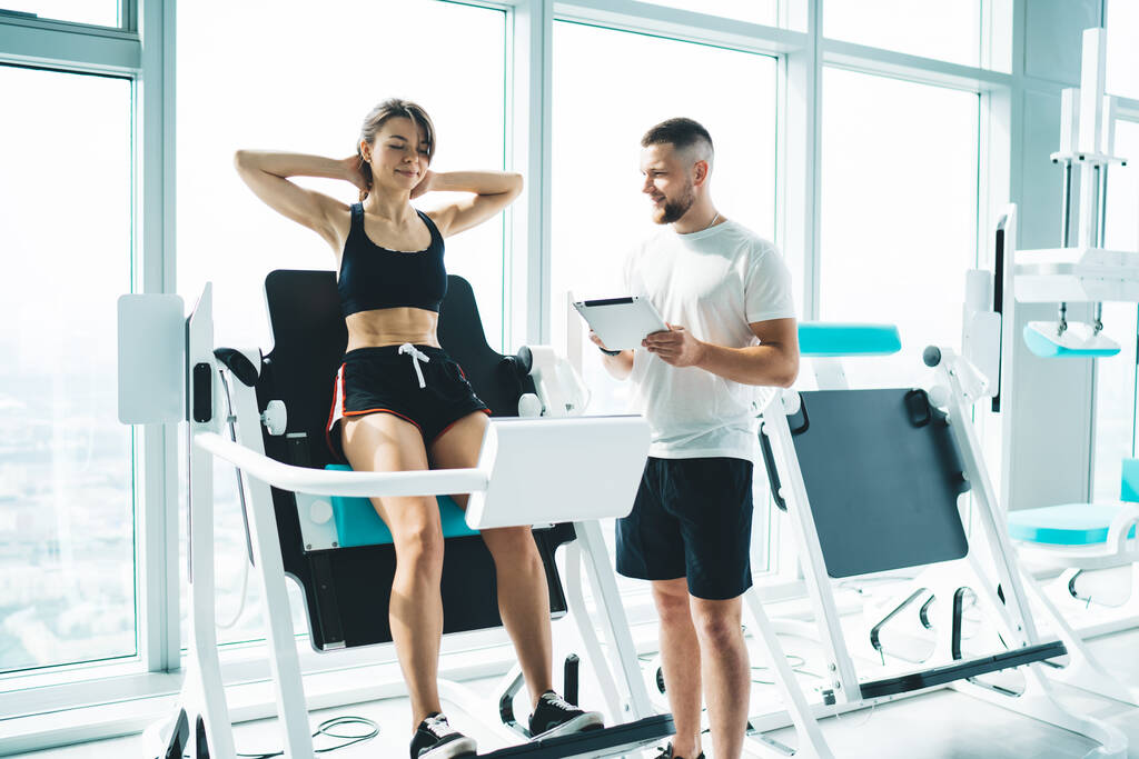 Junger fröhlicher männlicher Trainer mit Tablet interagiert mit fitter Frau beim Workout mit den Händen hinter dem Kopf auf digitalem Gerät im Fitnessstudio - Foto, Bild