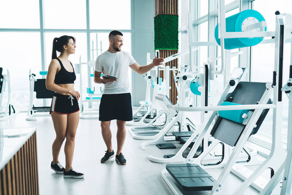 Full body of sportswoman and muscular coach wearing sportive wear walking in gym along modern fitness machines while choosing equipment for training - Photo, Image