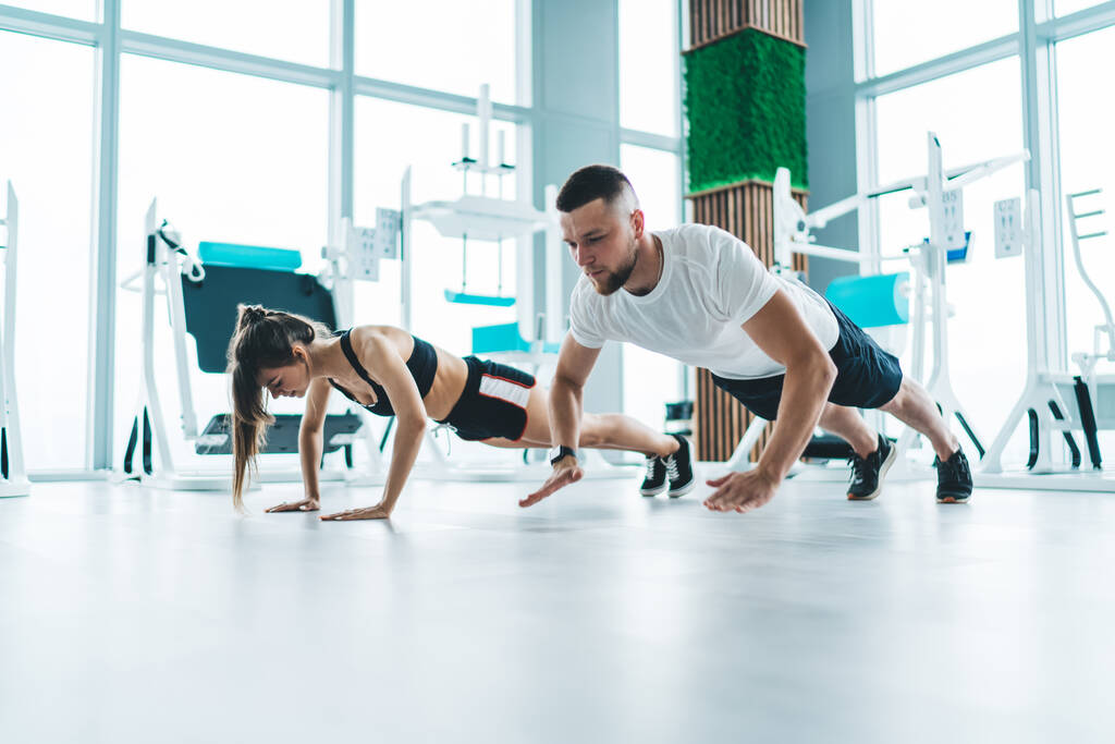 Повне тіло спортсмена і спортсменки в активному одязі роблять поштовхи на підлогу в сучасному тренажерному залі з великими світловими вікнами
 - Фото, зображення