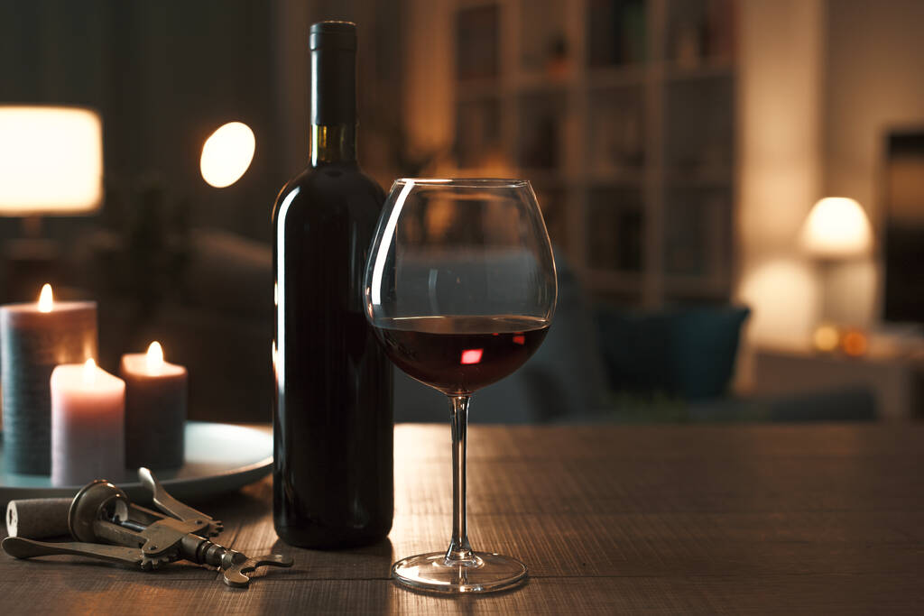 Ochutnávka červeného vína doma: láhev vína, sklenice na víno, vývrtka a svíčky na stole v obývacím pokoji v noci - Fotografie, Obrázek