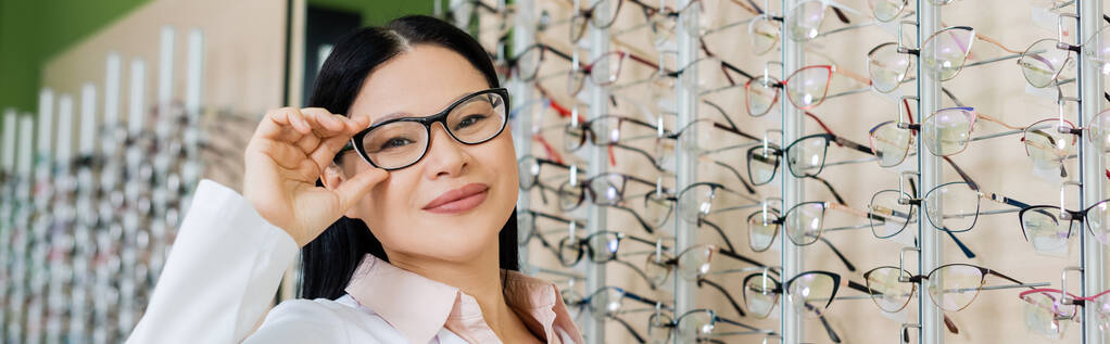 happy asian oculist looking at camera and touching eyeglasses in optics salon, banner - Fotó, kép